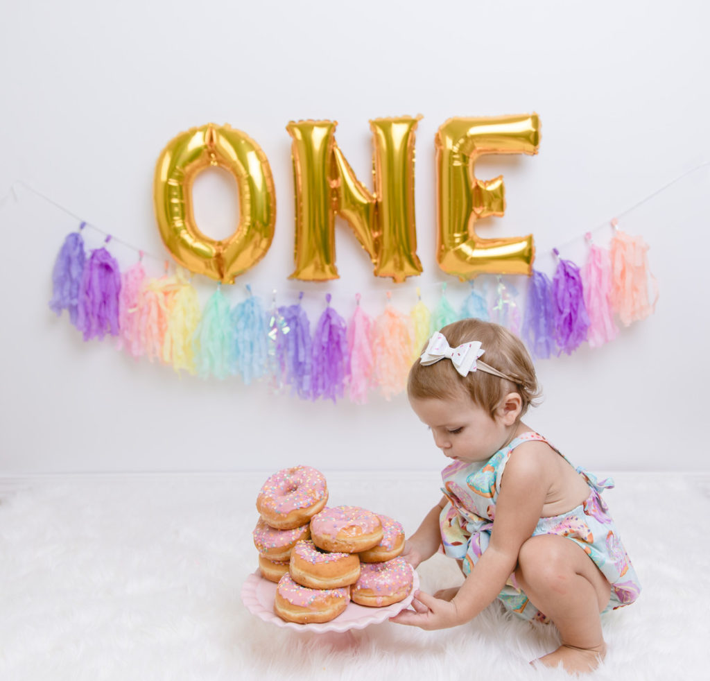 one year old girl grabbing cake stand of stacked pink frosted donuts at first birthday photoshoot