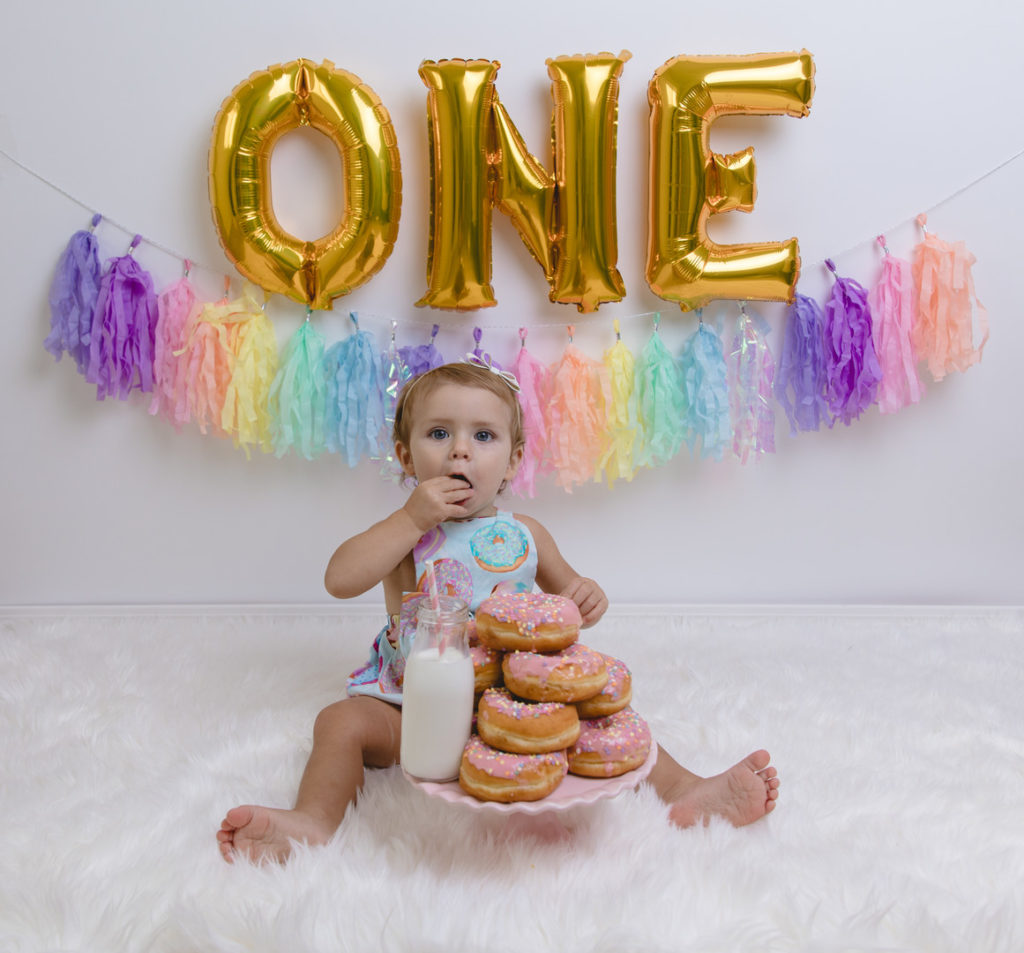 one year old girl eating pink donuts with a glass of milk, gold ONE banner on a white fur rug